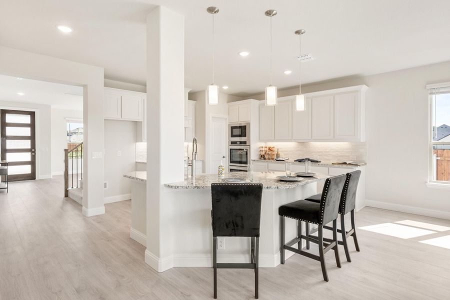 Open kitchen with white cabinets, granite island, black bar stools, and pendant lights over light wood floors