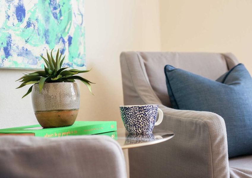 Staged home completed with two gray chairs one with blue pillow, small round table with vase with plant in it and blue cup beside, and turquoise colored painting hanging on the wall