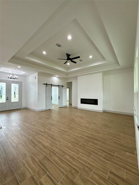This spacious living room features a high tray ceiling with recessed lighting and a modern fan. It has wood-look tile flooring, a sleek built-in fireplace, and a stylish barn door. Natural light enters through large windows and the entryway with double doors. This spacious living room features a high tray ceiling with recessed lighting and a modern fan. It has wood-look tile flooring, a sleek built-in fireplace, and a stylish barn door. Natural light enters through large windows and the entryway with double doors.