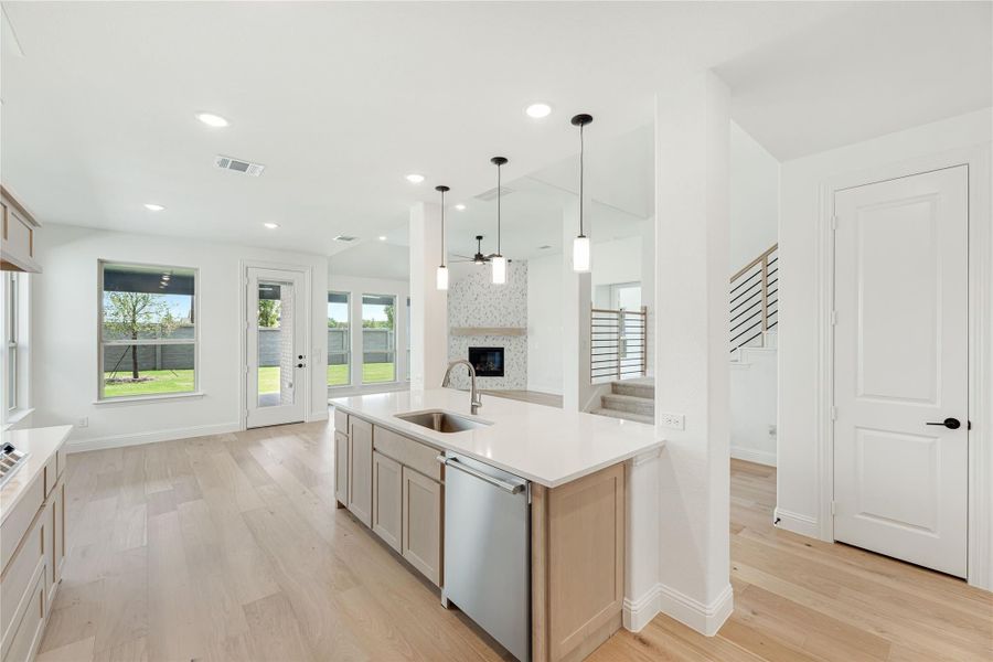 Kitchen island with sink and dishwasher, white countertops, pendant lights, and light wood floors open to living area with fireplace. Kitchen island with sink and dishwasher, white countertops, pendant lights, and light wood floors open to living area with fireplace.