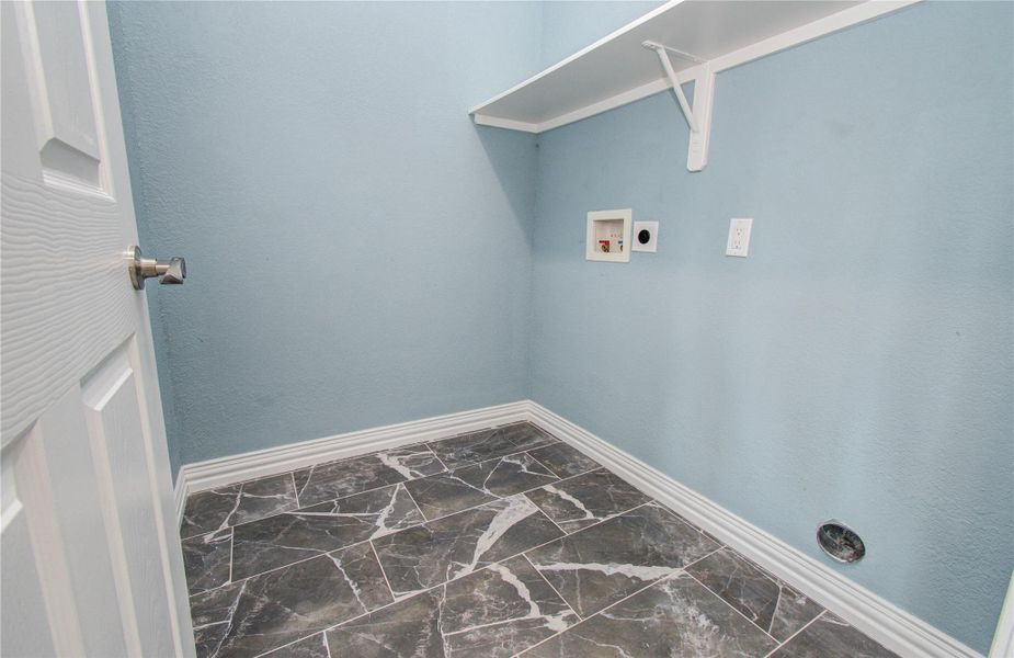 This photo shows a laundry room with light blue walls and dark marble-patterned tile flooring. It features a white shelf and hookups for a washer and dryer.