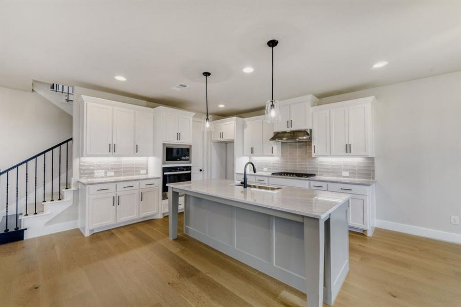 Contemporary kitchen featuring a central island with a sink, light-toned wood-finish flooring, white cabinetry with dark hardware, stainless steel appliances, and a tiled backsplash