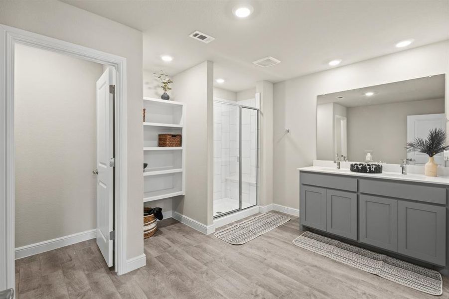 Bathroom featuring wood-finish flooring, a double vanity with gray cabinetry and white countertops, a glass-enclosed shower, and built-in shelving Bathroom featuring wood-finish flooring, a double vanity with gray cabinetry and white countertops, a glass-enclosed shower, and built-in shelving