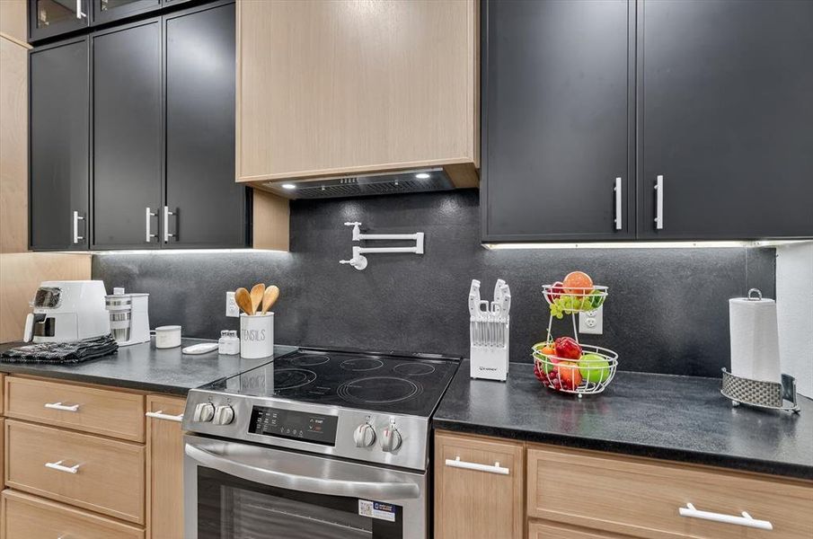 Modern kitchen featuring dual-tone cabinetry with light wood and dark finishes, stainless steel range with a smooth cooktop, pot filler faucet, dark countertops, and under-cabinet lighting