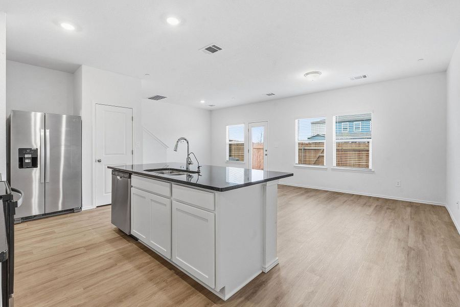 Open-concept living area featuring wood-finish flooring, a kitchen island with dark countertop and integrated sink, stainless steel appliances, and white cabinetry Open-concept living area featuring wood-finish flooring, a kitchen island with dark countertop and integrated sink, stainless steel appliances, and white cabinetry
