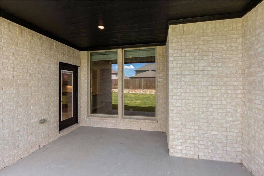 Covered back patio with light brick walls, dark ceiling, glass door, and large window overlooking fenced backyard