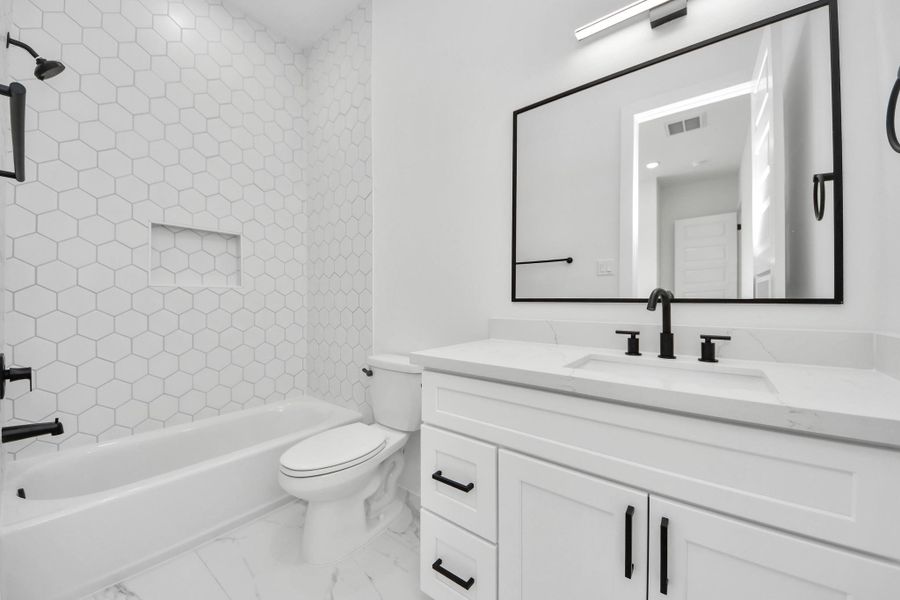 This modern bathroom features a sleek design with white hexagonal tile walls, a built-in bathtub, and contemporary black fixtures. The vanity offers ample storage and a large, framed mirror enhances the space.