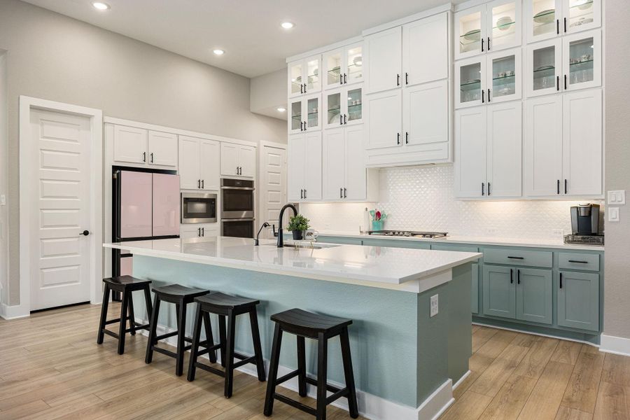 This bright and modern kitchen showcases an elegant design with tall white cabinetry, glass-front uppers, and sleek black hardware. A spacious island with quartz countertops provides ample workspace and seating, while the stylish two-tone cabinets add a touch of charm. Recessed lighting, a designer backsplash, and stainless steel built-in appliances complete this sophisticated culinary space.