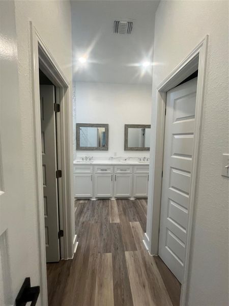 This photo showcases a modern bathroom with dual sinks, white cabinetry, and a clean, bright aesthetic. It features medium gray/ wood waterproof plank flooring and recessed lighting, creating a spacious and inviting atmosphere. Includes his and her walk-in-closets.