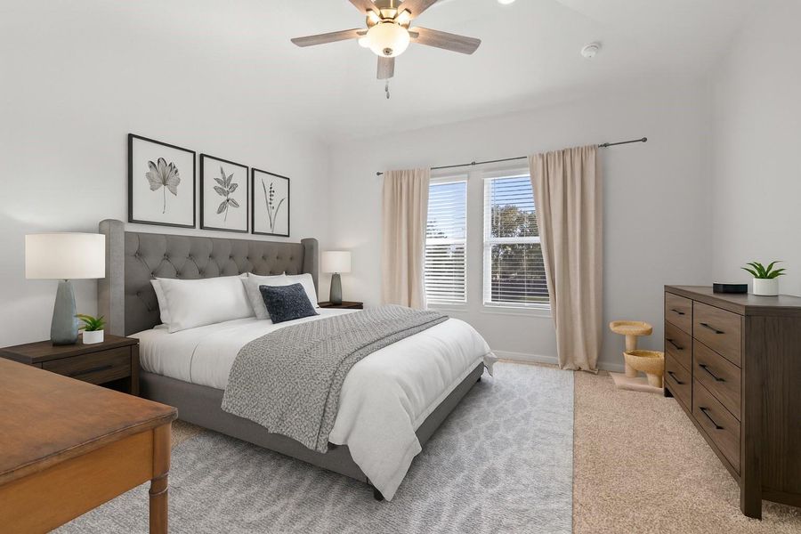 This bedroom features a cozy, modern design with a plush gray headboard, soft bedding, and neutral decor. Natural light fills the space through large windows, complemented by elegant curtains. The room includes a ceiling fan, two bedside tables with lamps, a dresser, and a small desk, creating a welcoming and functional environment.