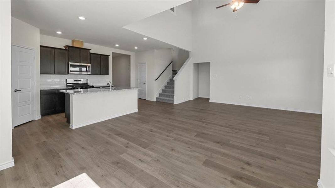 Open-concept living space featuring wood-finish flooring, a kitchen island with granite-style countertop, stainless steel appliances, dark cabinetry, and high ceilings