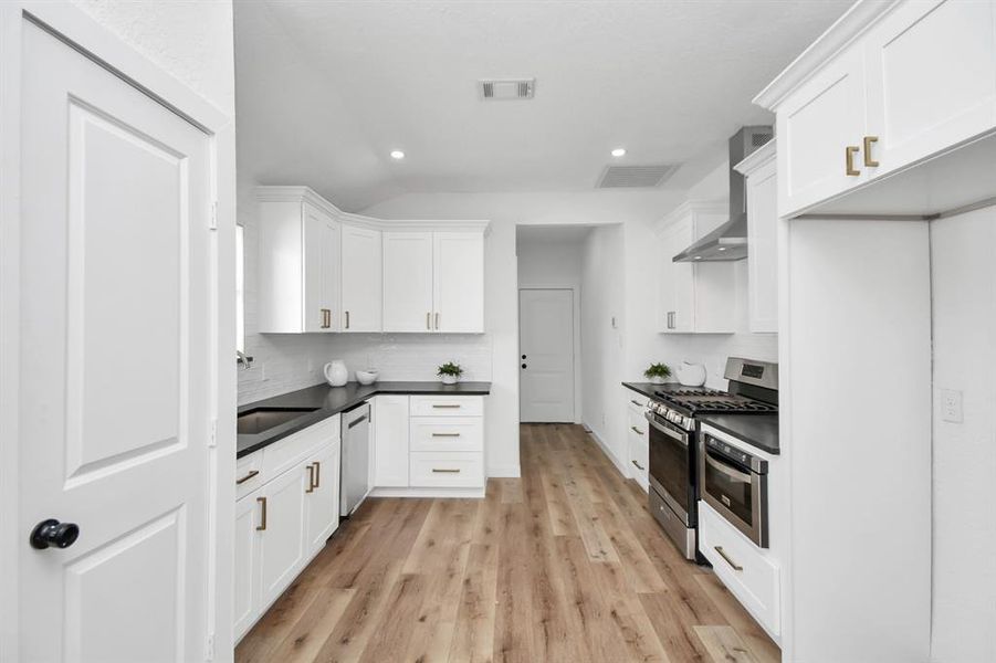 This is a modern, bright kitchen featuring white cabinetry, stainless steel appliances, a gas range, and a stylish subway tile backsplash. It has a sleek hood vent and appears to have ample storage space. The flooring is a light hardwood, and there's a subtle, minimalist aesthetic throughout.