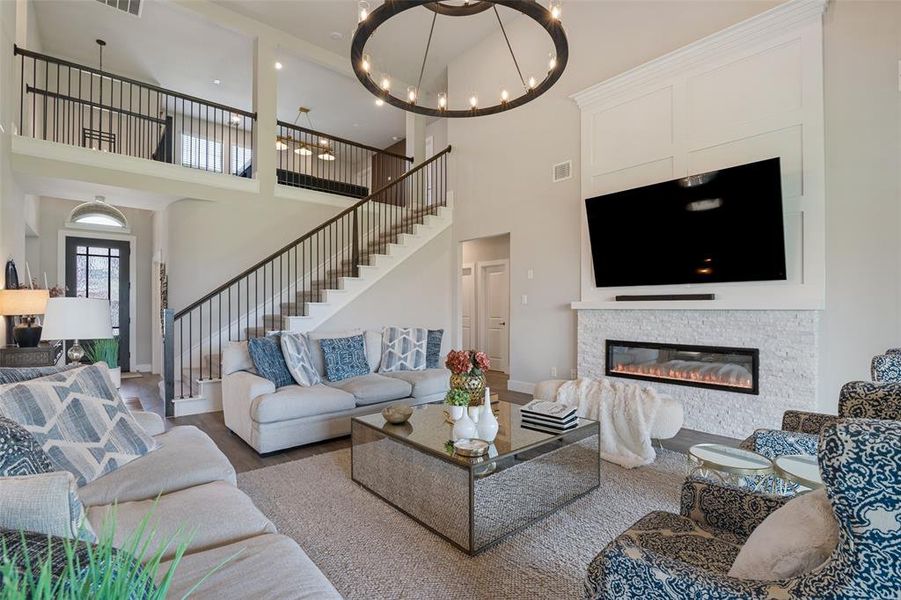 Two-story living room featuring a grand staircase with dark iron balusters, a built-in electric fireplace with a white stone surround, a large chandelier, and wood-finish flooring