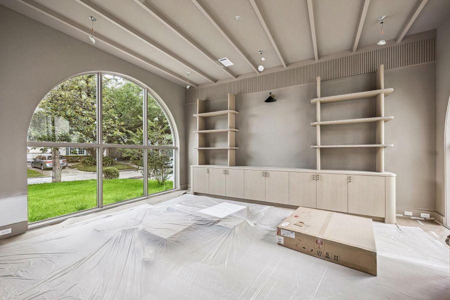 Library off the entrance of the home. This window is a dramatic architectural focal point, designed in a grand arched (radius top) configuration that immediately draws the eye. The Library features white-oak open shelving and built-in white oak cabinetry with flat panel doors.. The ceiling has painted beams and there are windows on both the north and south side of the room.  Construction as of June 1st, 2026.
