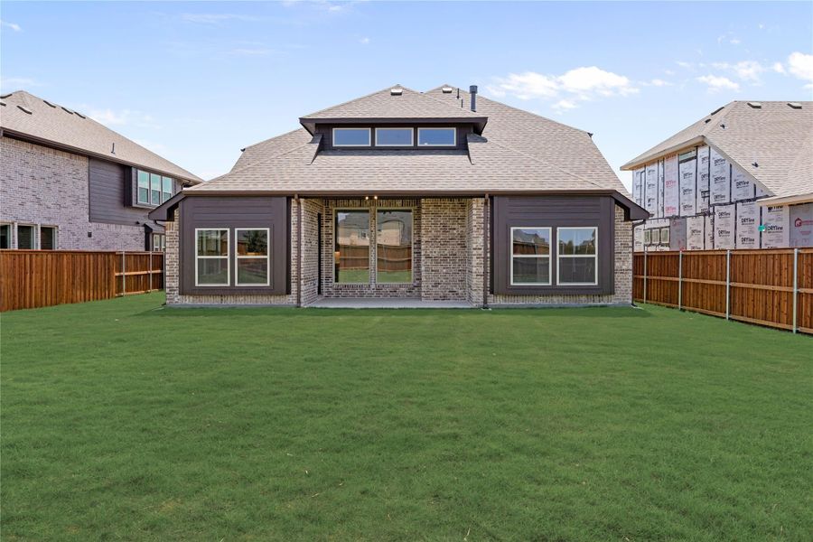 Rear exterior of brick and dark siding home with large windows, covered patio, and fenced backyard with green lawn