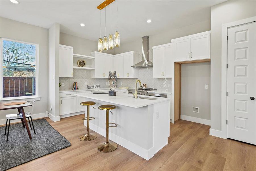 Kitchen with light countertops, hanging light fixtures, a kitchen island with sink, white cabinets, and wall chimney exhaust hood