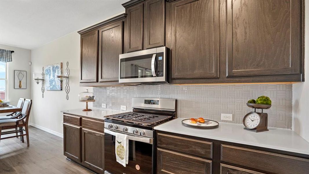 Kitchen featuring dark wood-finish cabinetry, stainless steel appliances, a gas range with an overhead microwave, white countertops, and a light-toned patterned tile backsplash Kitchen featuring dark wood-finish cabinetry, stainless steel appliances, a gas range with an overhead microwave, white countertops, and a light-toned patterned tile backsplash