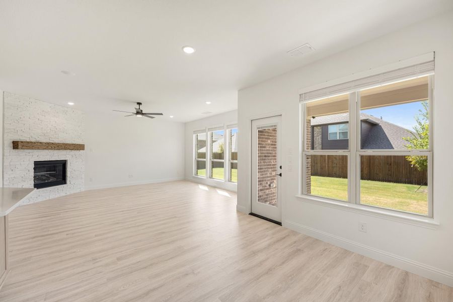 Open living room with stone fireplace, light wood floors, ceiling fan, and large windows overlooking backyard