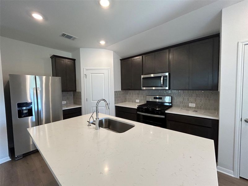 Kitchen featuring backsplash, appliances with stainless steel finishes, dark wood-style flooring, light stone counters, and an island with sink Kitchen featuring backsplash, appliances with stainless steel finishes, dark wood-style flooring, light stone counters, and an island with sink