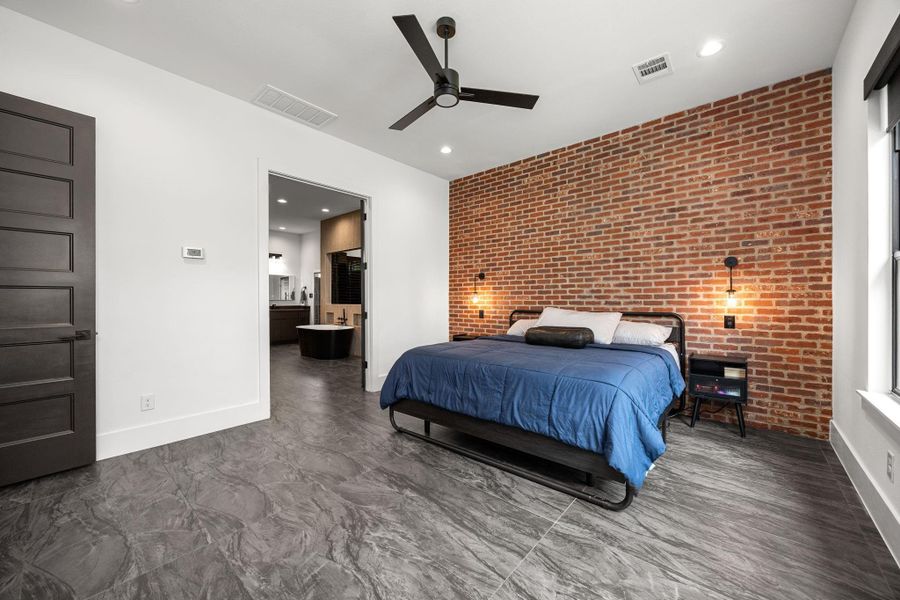 This modern bedroom features a striking exposed brick accent wall and sleek gray tile flooring. It includes a ceiling fan, recessed lighting, and direct access to a contemporary bathroom with a freestanding tub. This modern bedroom features a striking exposed brick accent wall and sleek gray tile flooring. It includes a ceiling fan, recessed lighting, and direct access to a contemporary bathroom with a freestanding tub.