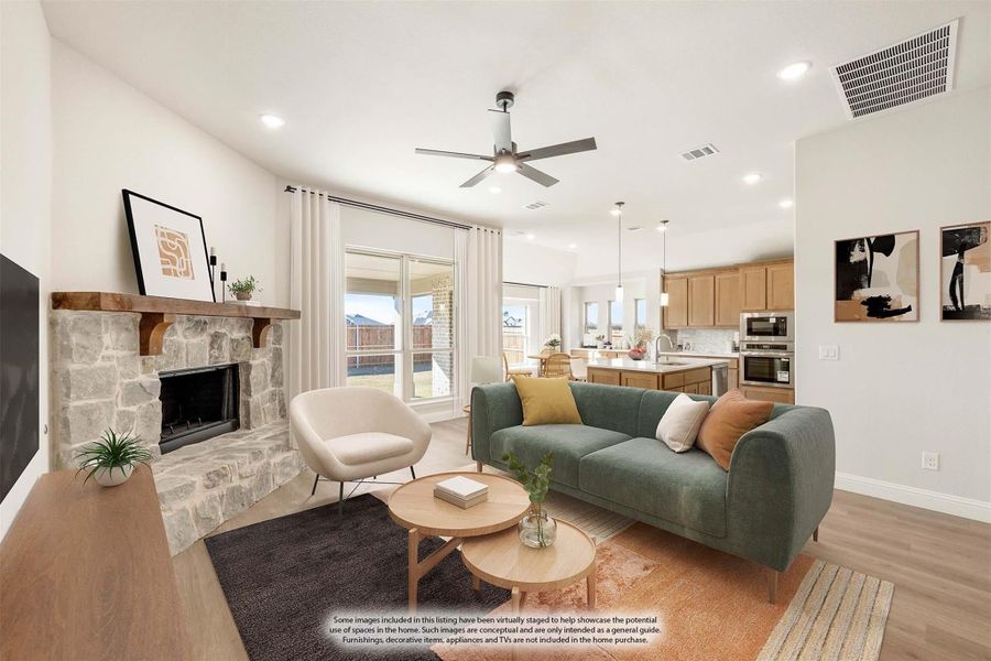 Open-concept living room with stone fireplace, ceiling fan, and view into kitchen with wood cabinets