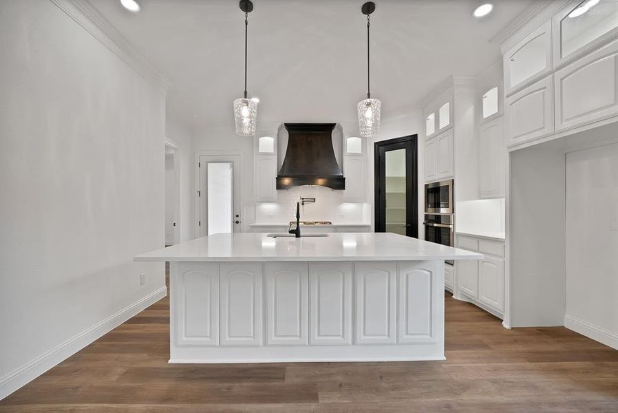 Kitchen with white cabinets, decorative light fixtures, dark wood-style flooring, premium range hood, and an island with sink