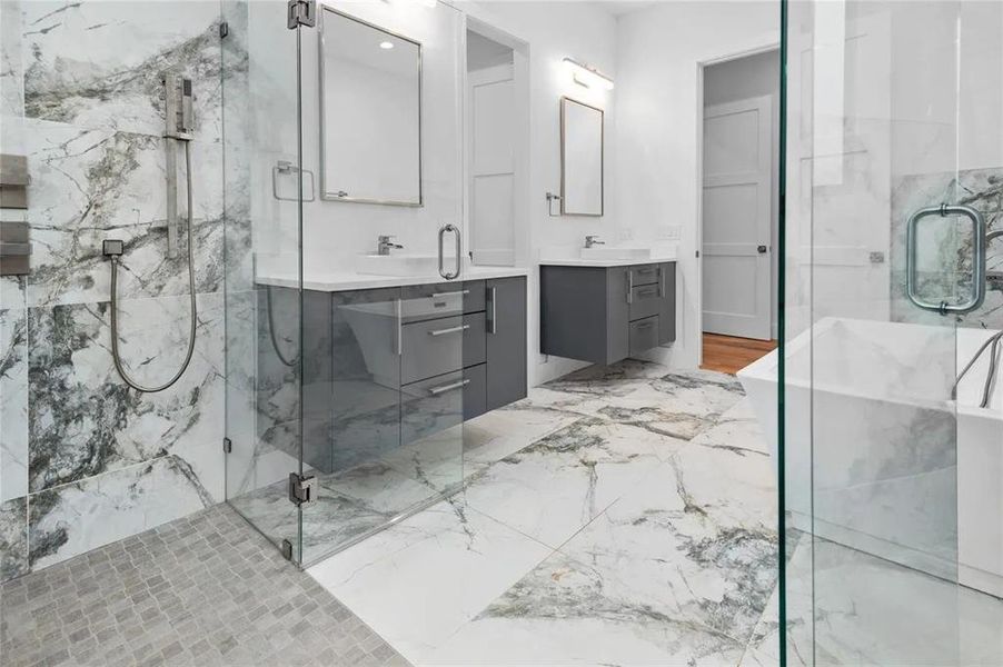 Modern bathroom featuring a glass-enclosed shower with a mosaic tile floor, a freestanding soaking tub, and a dual vanity with grey cabinetry Modern bathroom featuring a glass-enclosed shower with a mosaic tile floor, a freestanding soaking tub, and a dual vanity with grey cabinetry