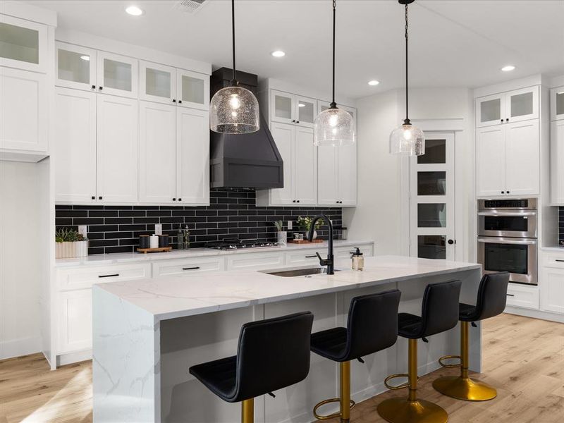 Kitchen featuring a waterfall island with an undermount sink, white cabinetry with black hardware, a black subway tile backsplash, a black range hood, and integrated stainless steel double ovens