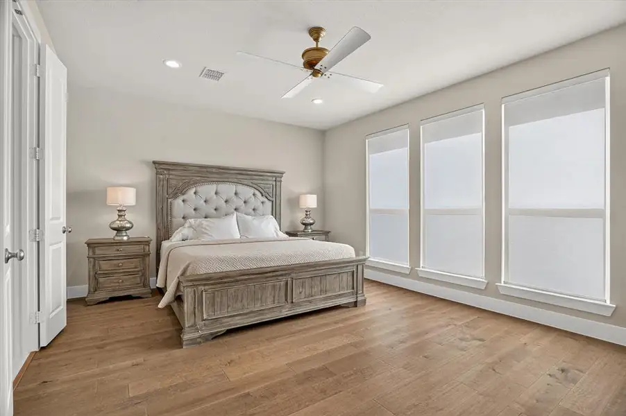 Spacious room featuring wood-finish flooring, three vertical windows with roller shades, and a ceiling fan with integrated lighting Spacious room featuring wood-finish flooring, three vertical windows with roller shades, and a ceiling fan with integrated lighting