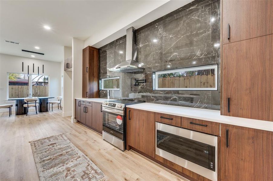 Kitchen with stainless steel appliances, wall chimney range hood, tasteful backsplash, light wood-type flooring, and brown cabinets