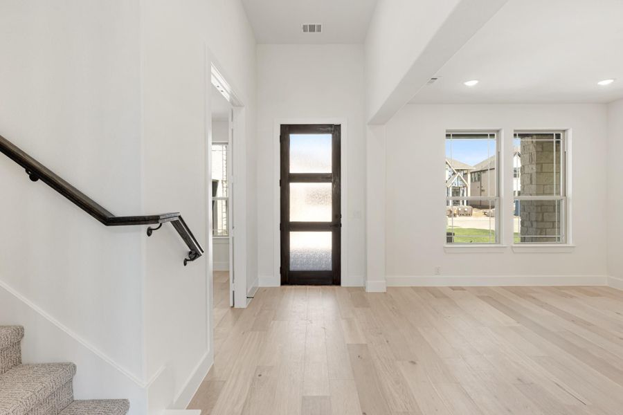 Bright entryway with light hardwood floors, dark glass-panel door, staircase, and white walls