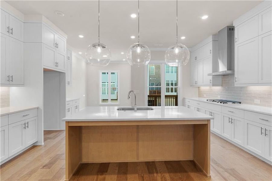 Kitchen featuring tasteful backsplash, white cabinetry, hanging light fixtures, wall chimney exhaust hood, and light wood-style flooring