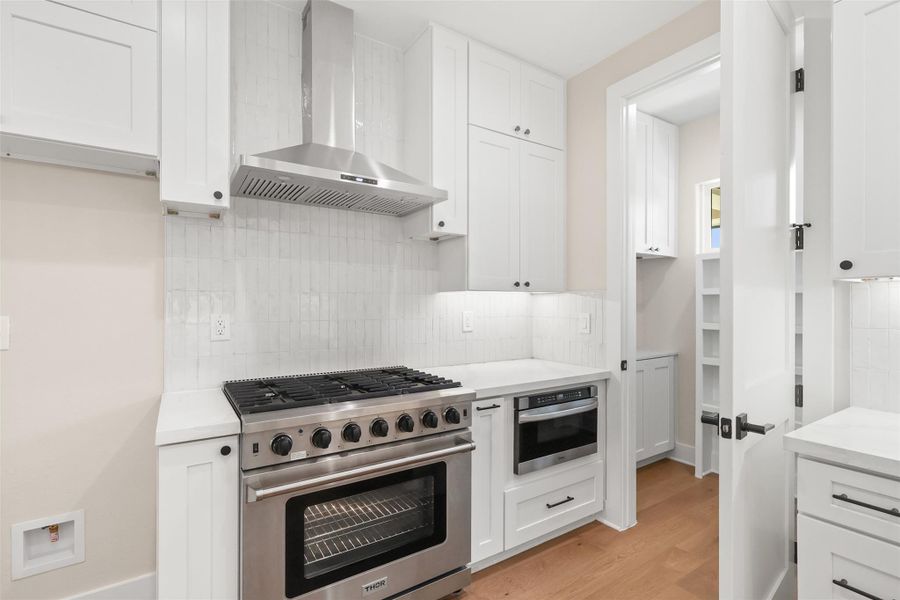 Kitchen featuring tasteful backsplash, stainless steel appliances, light countertops, wall chimney range hood, and light wood-type flooring Kitchen featuring tasteful backsplash, stainless steel appliances, light countertops, wall chimney range hood, and light wood-type flooring