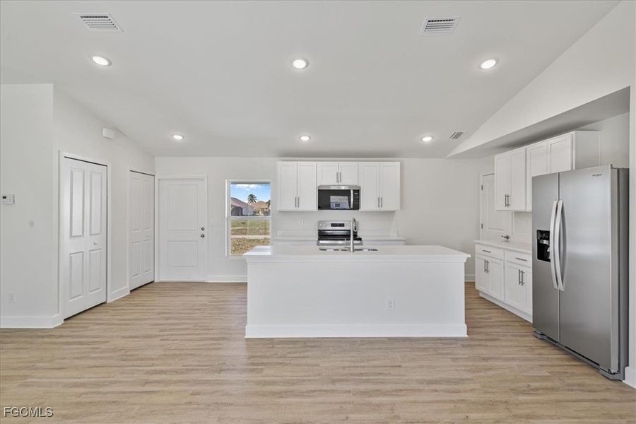 Kitchen featuring appliances with stainless steel finishes, vaulted ceiling, white cabinetry, an island with sink, and light wood-type flooring