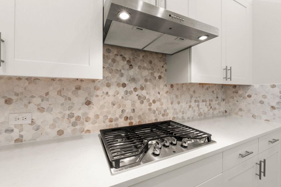 Kitchen with ventilation hood, stainless steel gas stovetop, light countertops, white cabinets, and decorative backsplash Kitchen with ventilation hood, stainless steel gas stovetop, light countertops, white cabinets, and decorative backsplash