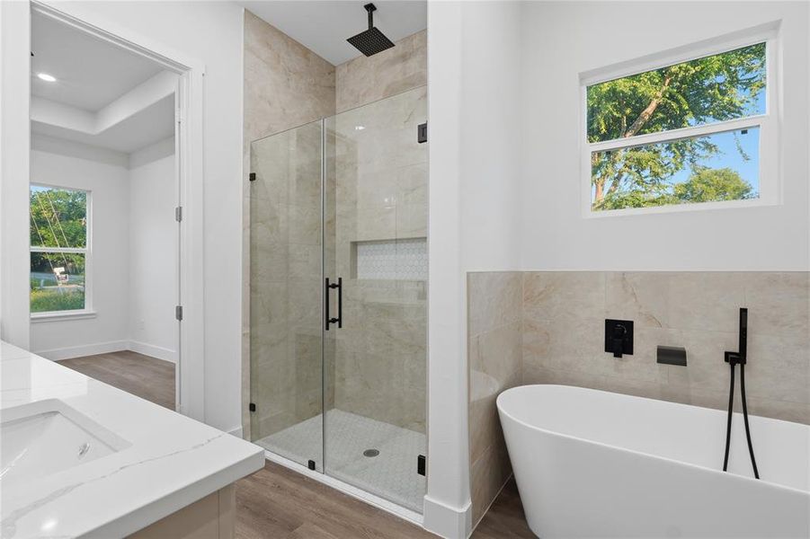 Full-glass shower enclosure with a rain shower head, freestanding soaking tub with wall-mounted matte black fixtures, quartz countertop vanity, wood-finish flooring, and a large window