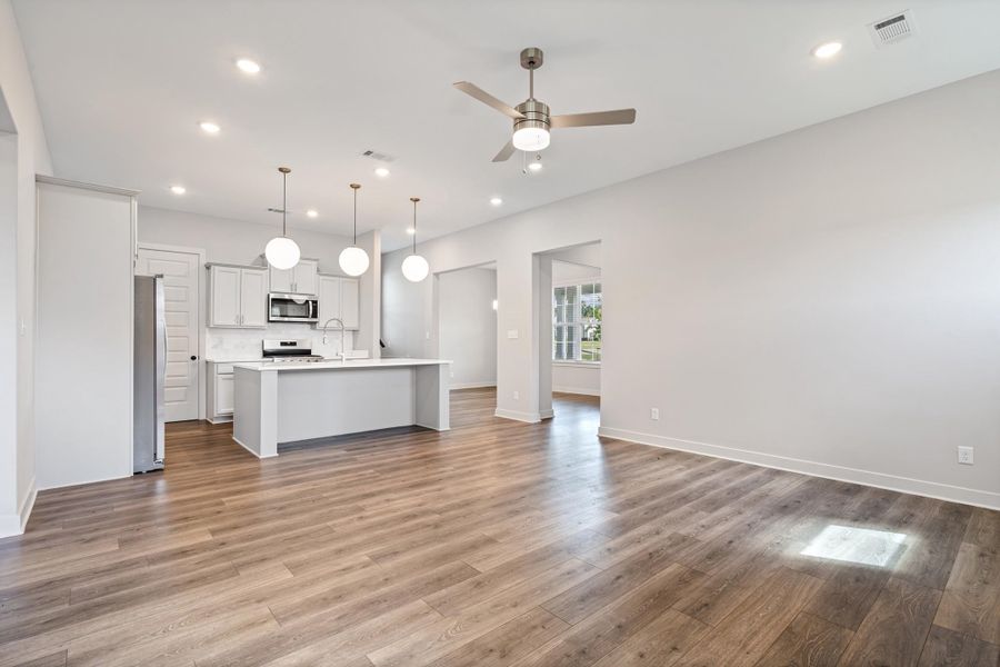 Open-concept living and kitchen area featuring hardwood flooring throughout, a white cabinetry kitchen island with stainless steel appliances, recessed and pendant lighting, a ceiling fan, and abundant natural light from multiple windows