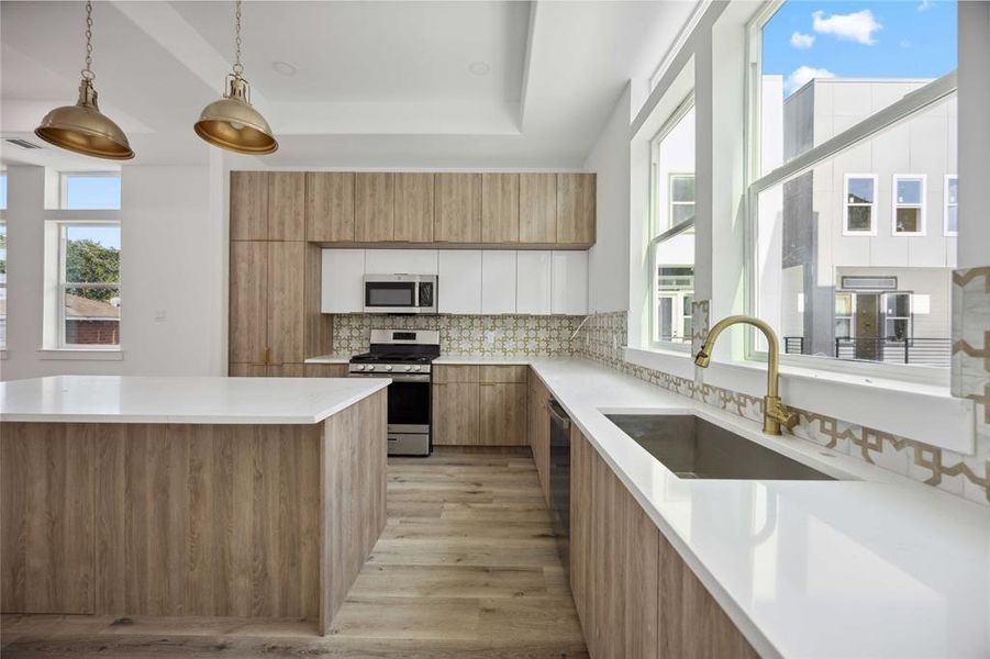 Modern kitchen featuring light cabinetry, sleek white countertops, stainless steel appliances, and pendant lighting, accented by a decorative backsplash. Large windows allow for ample natural light!