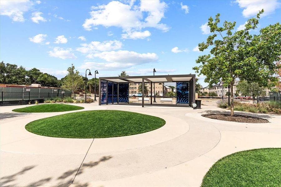 Community gathering area featuring a modern pergola structure, curvilinear paved pathways, two mounded turf islands, and established trees Community gathering area featuring a modern pergola structure, curvilinear paved pathways, two mounded turf islands, and established trees