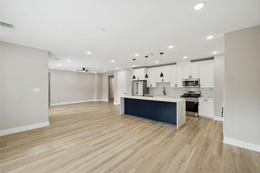 Open-concept living space with light wood flooring and recessed lighting. The kitchen features white cabinetry, a large island with a dark blue base, stainless steel appliances, and a subway tile backsplash.