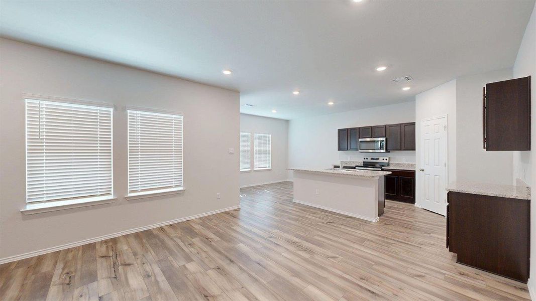 Spacious open-concept living area featuring wood-finish flooring, recessed lighting, multiple windows with blinds, a kitchen island, and dark wood cabinetry with light countertops Spacious open-concept living area featuring wood-finish flooring, recessed lighting, multiple windows with blinds, a kitchen island, and dark wood cabinetry with light countertops