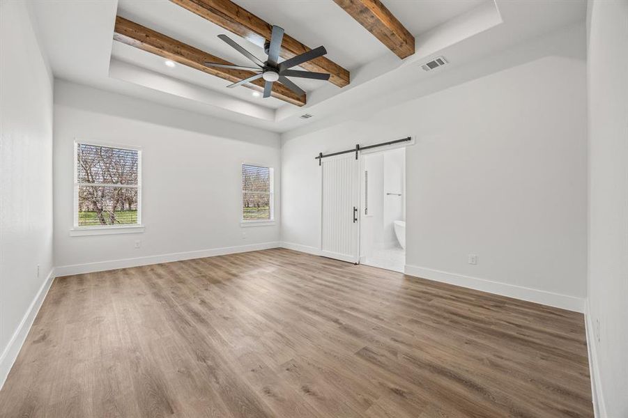Unfurnished bedroom featuring beamed ceiling, a barn door, a raised ceiling, wood finished floors, and recessed lighting Unfurnished bedroom featuring beamed ceiling, a barn door, a raised ceiling, wood finished floors, and recessed lighting