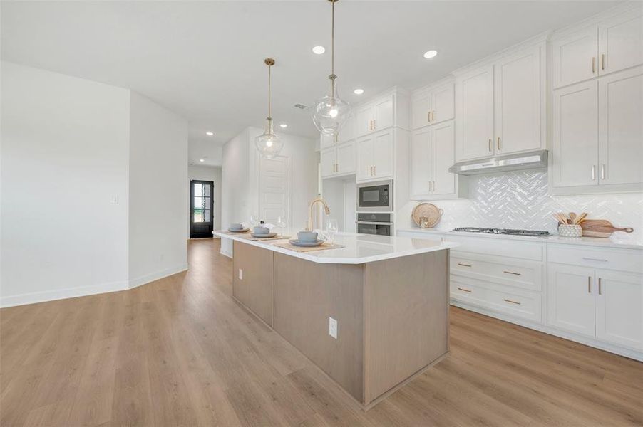 Open-concept kitchen featuring an island, light wood-finish flooring, white cabinetry, a herringbone tile backsplash, and stainless steel appliances