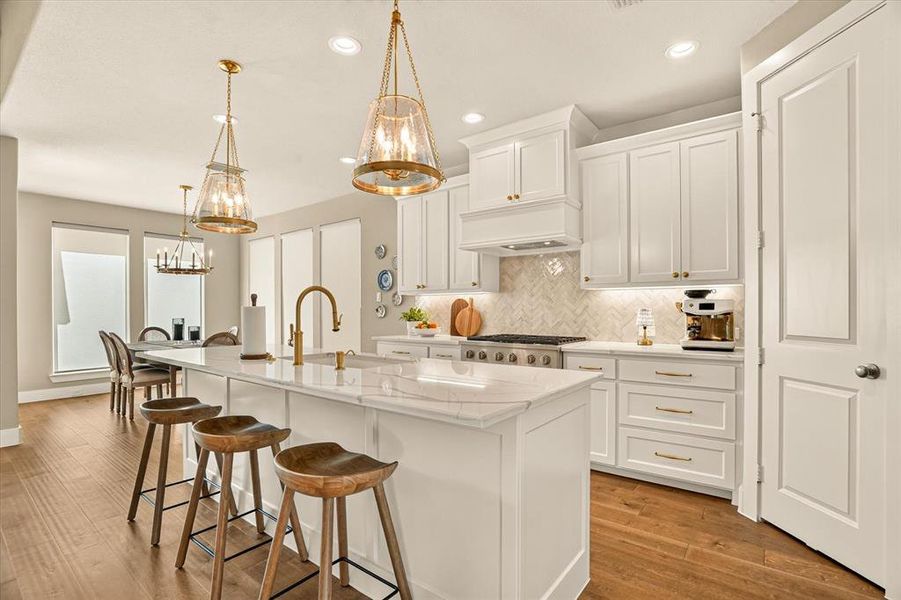 Open-concept kitchen featuring an oversized island with countertop seating, white shaker cabinetry, gold-finish hardware, and wood-finish flooring Open-concept kitchen featuring an oversized island with countertop seating, white shaker cabinetry, gold-finish hardware, and wood-finish flooring