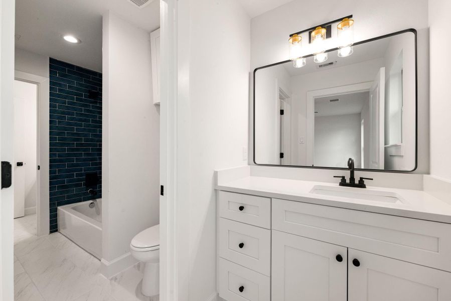 Modern bathroom featuring a sleek white vanity with quartz countertop, black hardware, and a large framed mirror with contemporary lighting. The space is complemented by stylish tile flooring and a tub/shower combo with striking dark tile accent for a bold, modern touch.