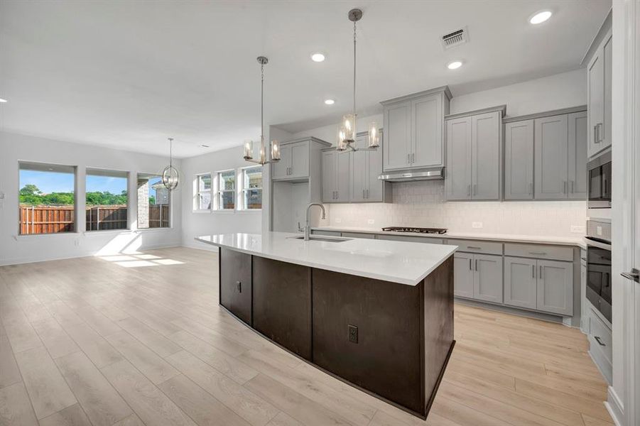 Kitchen featuring a chandelier, gray cabinetry, a kitchen island with sink, stainless steel appliances, and light wood-style floors Kitchen featuring a chandelier, gray cabinetry, a kitchen island with sink, stainless steel appliances, and light wood-style floors