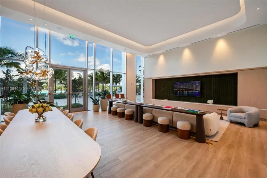 Floor-to-ceiling glass walls, wood-finish flooring, recessed tray ceiling with cove lighting, custom built-in media console, and statement multi-pendant lighting fixture