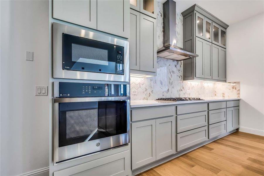 Kitchen with built in microwave, gray cabinetry, gas cooktop, light wood-type flooring, and wall chimney exhaust hood