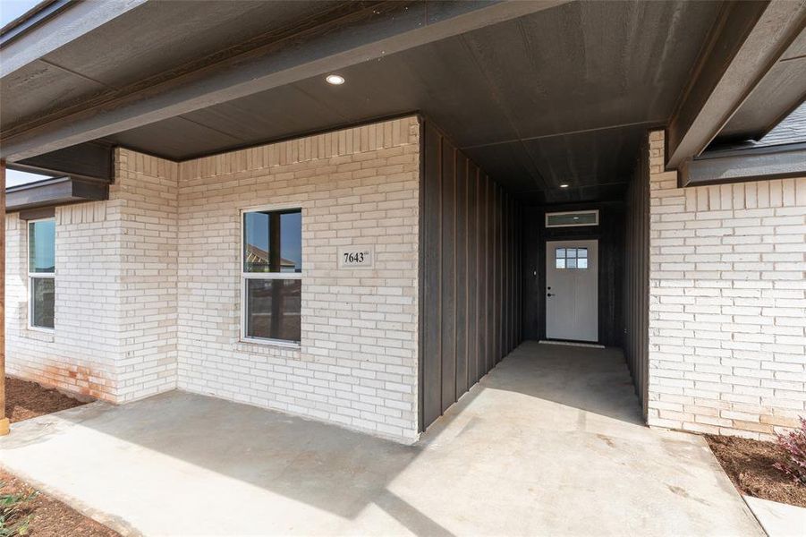 Covered entry featuring light brick facade, dark wood-finish siding, recessed lighting, concrete walkway, and a paneled front door with decorative glass inserts