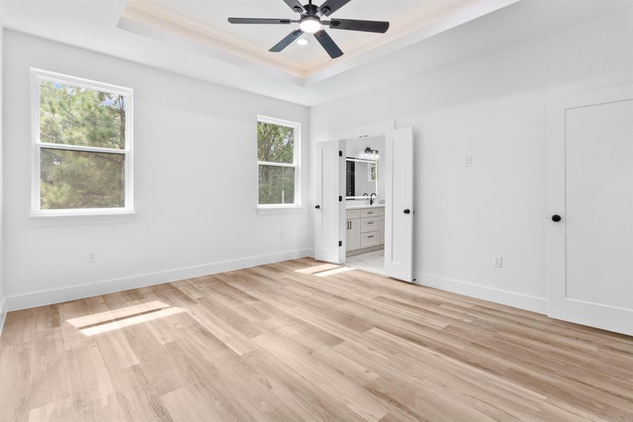 Unfurnished bedroom with ensuite bathroom, light wood-style floors, a ceiling fan, and a raised ceiling